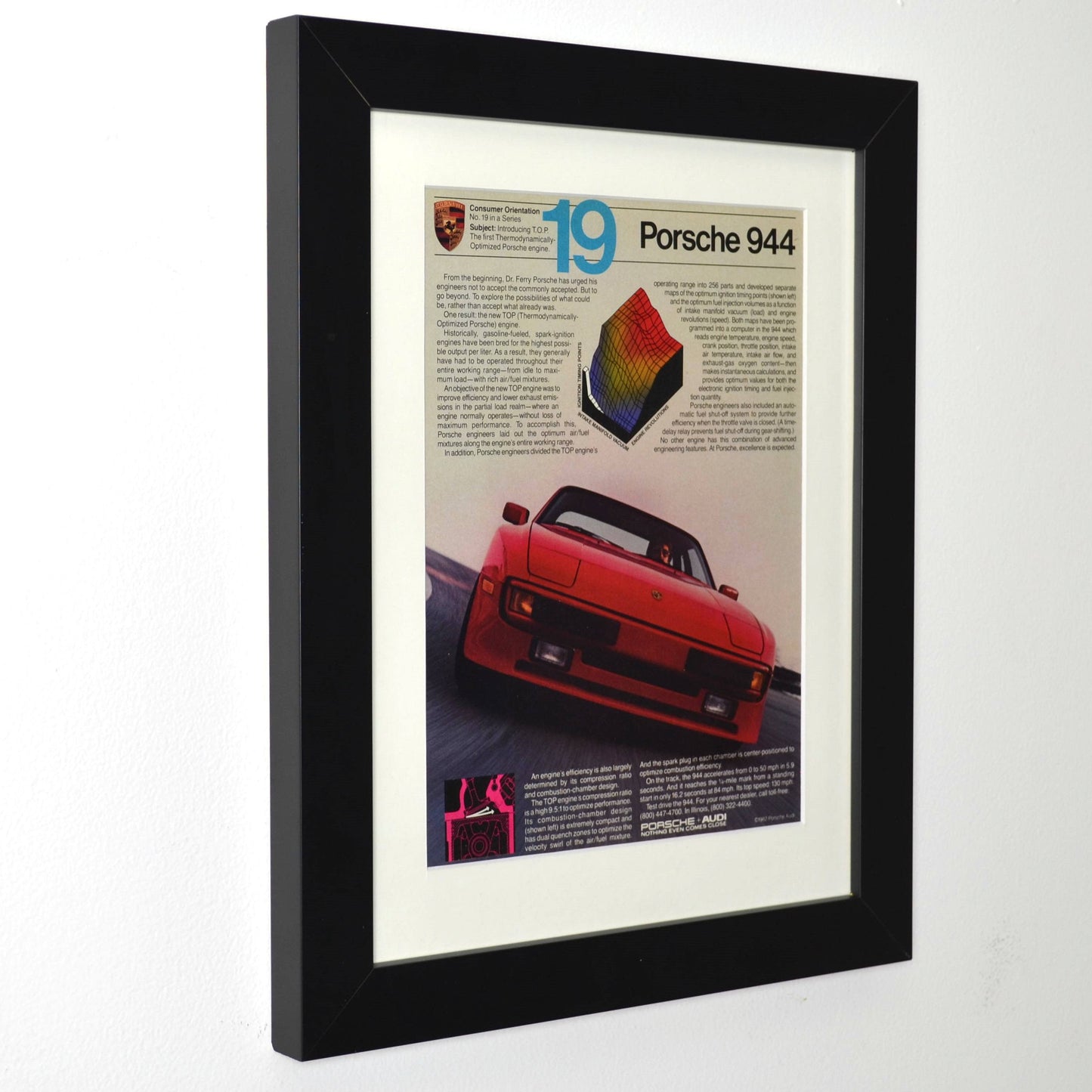 Framed vintage 1982 Porsche 944 magazine ad featuring red 944 in motion with technical graphics, dry-mounted and matted in black frame for wall display side view.