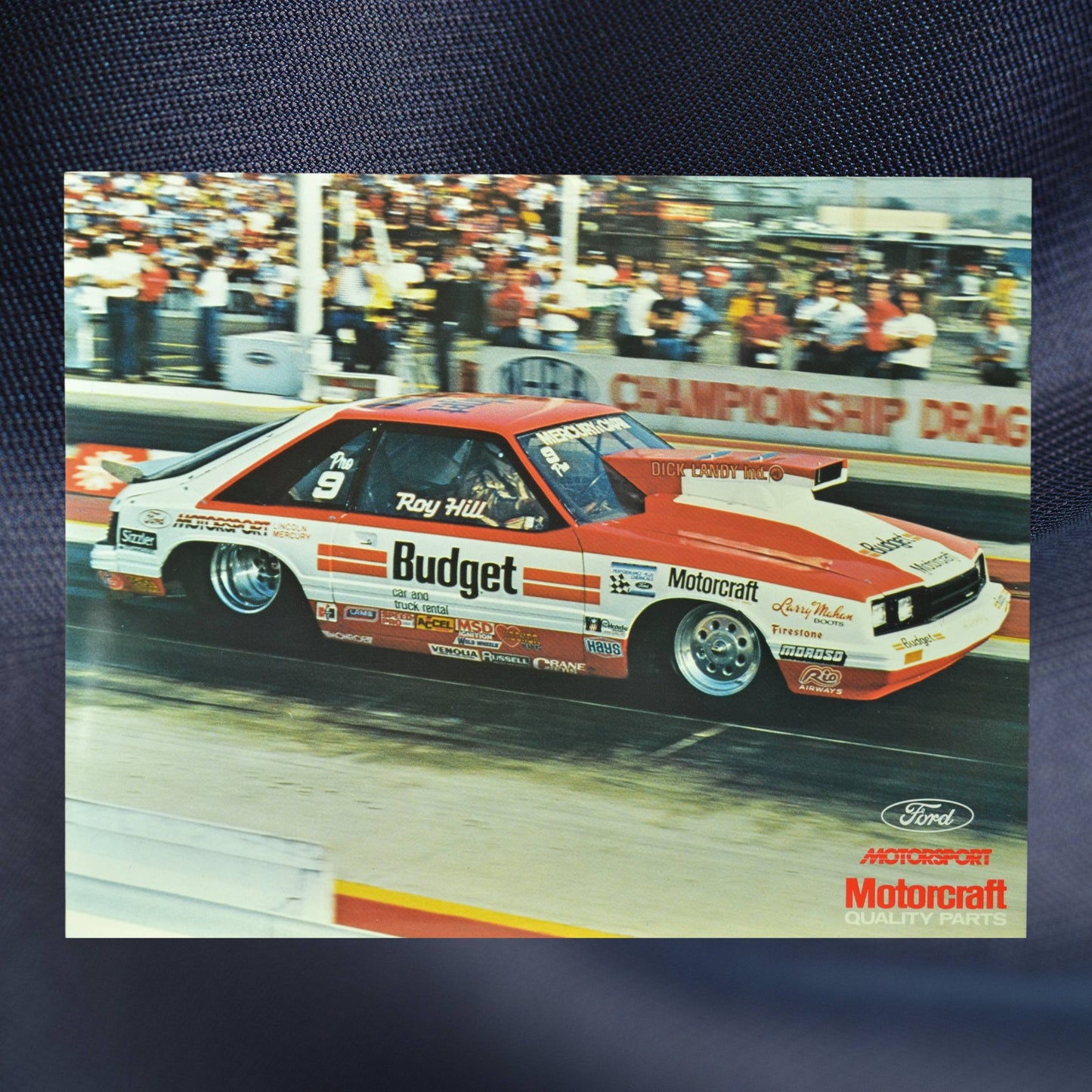 1984 NHRA hero card showing Roy Hill’s Budget-sponsored Mercury Capri Pro Stock car launching down the dragstrip with a crowd in the background.