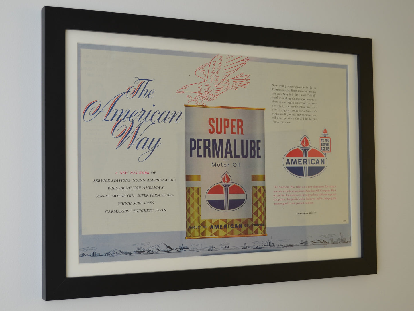 961 American Oil Super Permalube framed vintage print featuring a patriotic-themed gas and oil advertisement with the classic American Oil torch logo, dry-mounted and displayed in a black wood frame with a white matte. Ideal petroliana wall art for collectors and vintage gas station memorabilia enthusiasts, side view