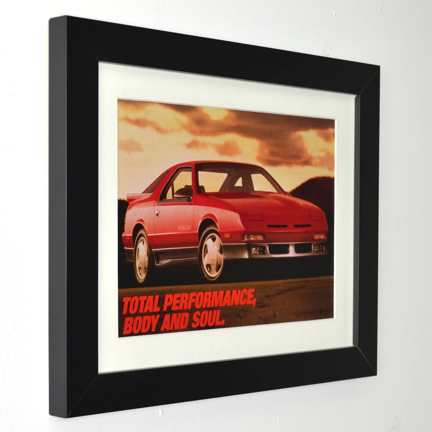 Framed 1989 Dodge Daytona Shelby vintage print ad featuring a red Daytona under a sunset sky with the slogan “Total Performance, Body and Soul.” Displayed in a matte black frame with white mat—authentic Mopar wall art for collectors side view