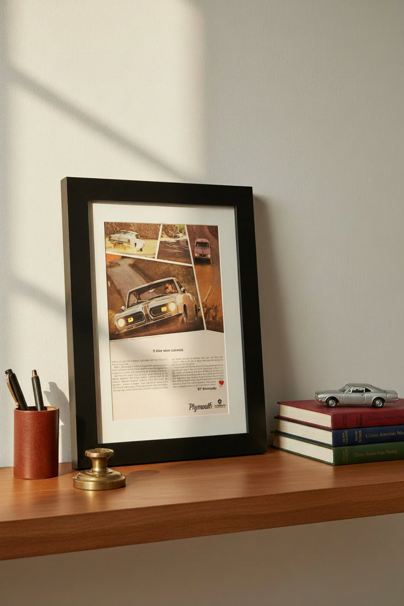 A framed vintage Plymouth Barracuda advertisement print, featuring a black wood frame and white mat board, showcasing a classic car on a road with printed text below.