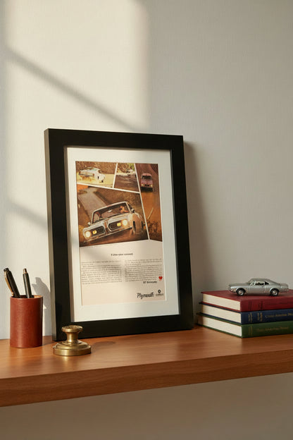 A framed vintage Plymouth Barracuda advertisement print, featuring a black wood frame and white mat board, showcasing a classic car on a road with printed text below.