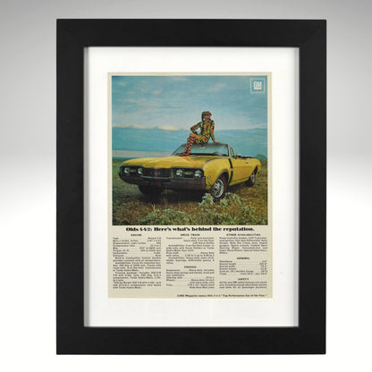 Framed 1968 Oldsmobile 442 convertible print ad – yellow 442 with woman sitting on hood in scenic field.