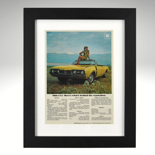 Framed 1968 Oldsmobile 442 convertible print ad – yellow 442 with woman sitting on hood in scenic field.