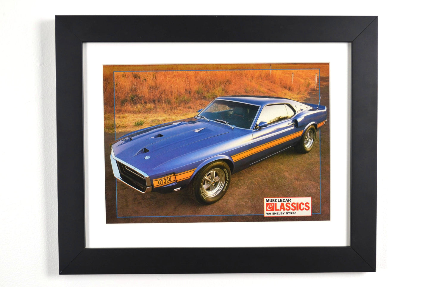 Framed 1969 Shelby GT-350 vintage print ad, featuring the bold blue Mustang with gold stripes, an iconic muscle car perfect for collectors and Ford fans.