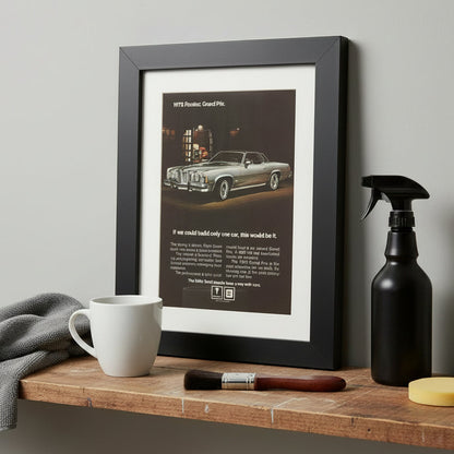 A framed vintage 1975 Pontiac Grand Prix advertisement print, featuring a black frame, white mat, and a dark brown ad with a silver convertible car and text.