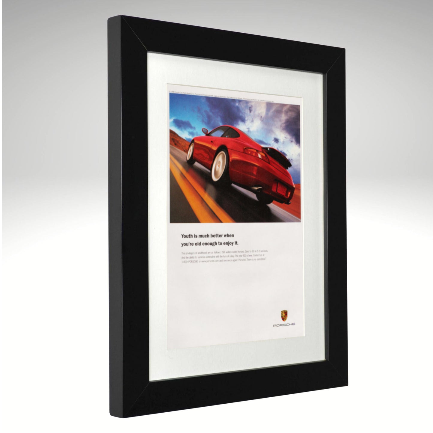 A framed Porsche print featuring a red sports car on a road with the text 'Youth is much better when you're well enough to control it.' and the Porsche crest at the bottom, set in a black frame with a white mat against a light gray background.