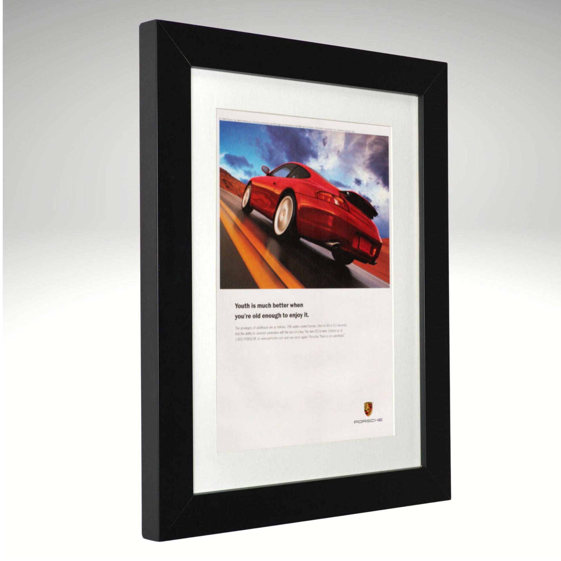 A framed Porsche print featuring a red sports car on a road with the text 'Youth is much better when you're well enough to control it.' and the Porsche crest at the bottom, set in a black frame with a white mat against a light gray background.