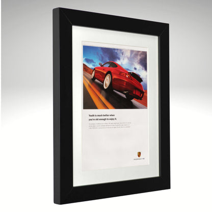 A framed Porsche print featuring a red sports car on a road with the text 'Youth is much better when you're well enough to control it.' and the Porsche crest at the bottom, set in a black frame with a white mat against a light gray background.