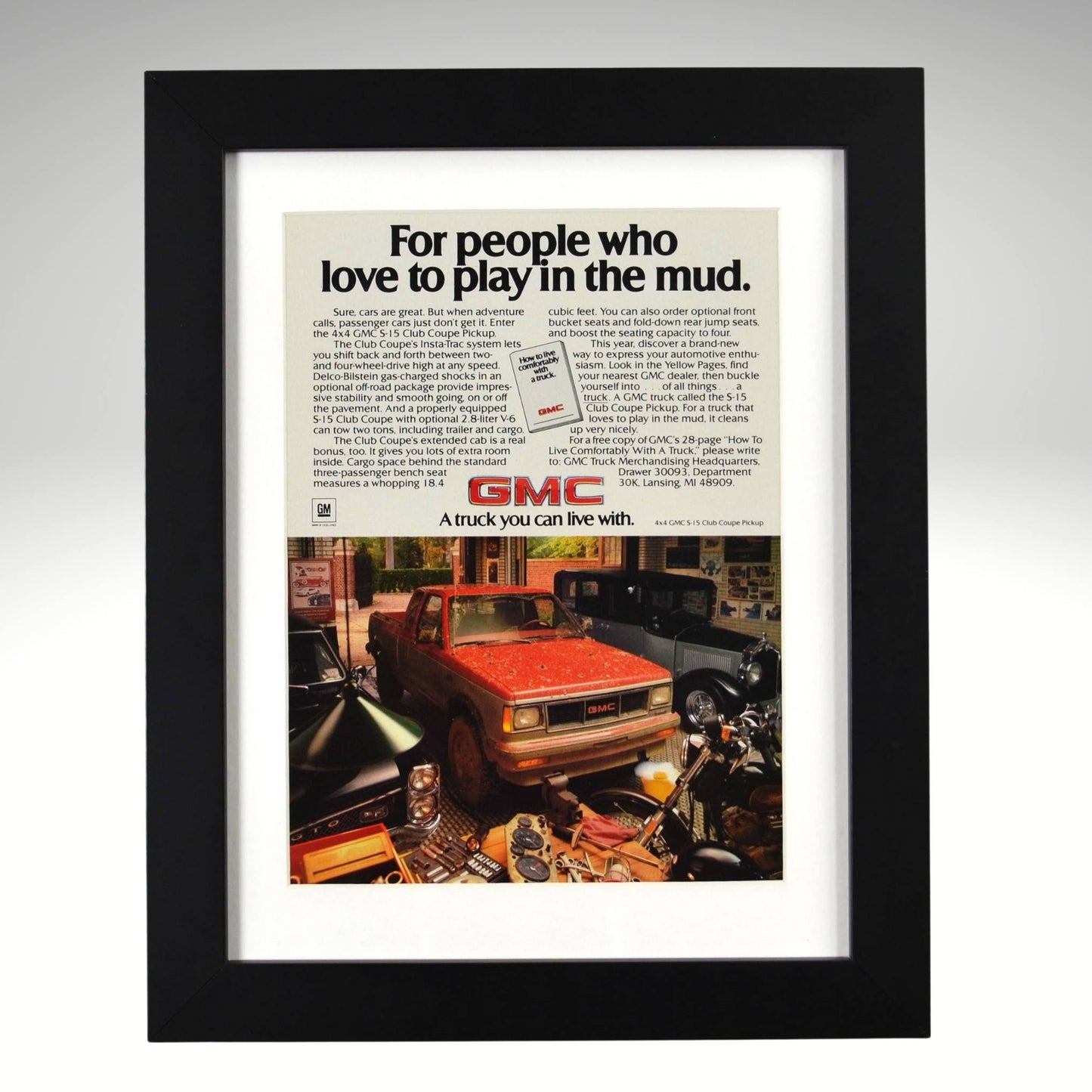 Framed GMC S-15 4x4 print ad – red compact pickup covered in mud, vintage garage scene with tools and motorcycles.

