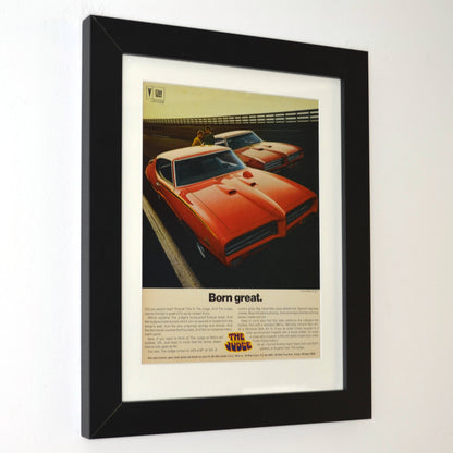Framed 1969 Pontiac GTO Judge vintage print ad featuring two Carousel Red GTOs with headline “Born Great.” Displayed in a matte black frame with white mat — classic muscle car wall art for collectors and enthusiasts side view