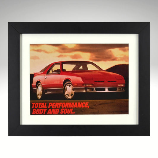 1989 Dodge Daytona Shelby vintage print ad featuring a red Daytona under a sunset sky with the slogan “Total Performance, Body and Soul.” Displayed in a matte black frame with white mat—authentic Mopar wall art for collectors.