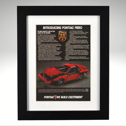 A framed vintage Pontiac Fiero advertisement print, featuring a red sports car and printed text, enclosed in a white mat and a black frame against a light gray background.