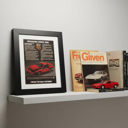 A framed vintage 1984 Pontiac Fiero advertisement print, featuring a black frame, white mat, and a red car graphic on a light gray gradient background.
