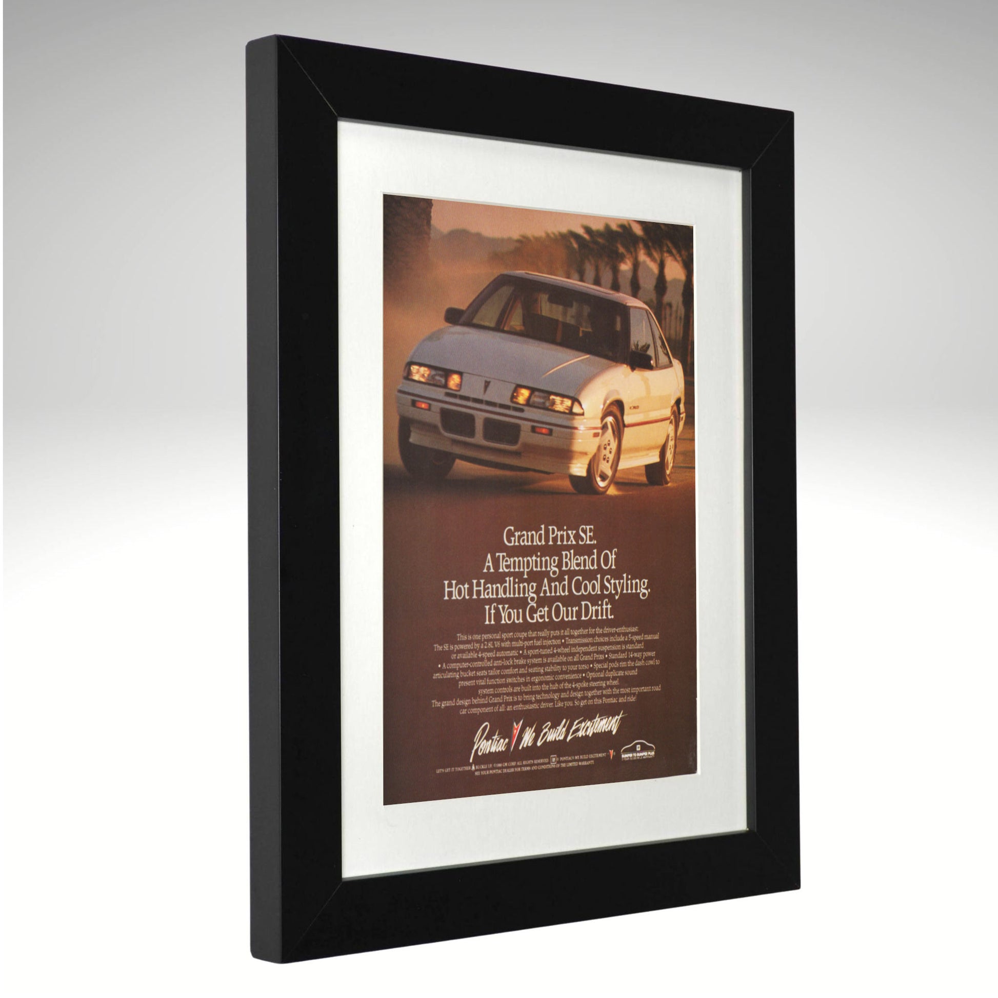 A framed vintage Pontiac Grand Prix SE advertisement print, featuring a white sports car on a warm sunset road with the headline 'Grand Prix SE' and smaller text below, set in a black frame with a white mat against a light gray background.