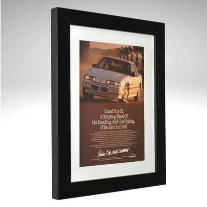 A framed vintage Pontiac Grand Prix SE advertisement print, featuring a white sports car on a warm sunset road with the headline 'Grand Prix SE' and smaller text below, set in a black frame with a white mat against a light gray background.