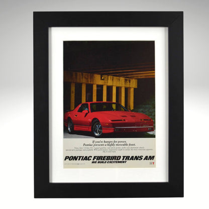 Framed 1986 Pontiac Firebird Trans Am WS6 ad – vintage print with red Trans Am under overpass, 1980s performance car wall art.

