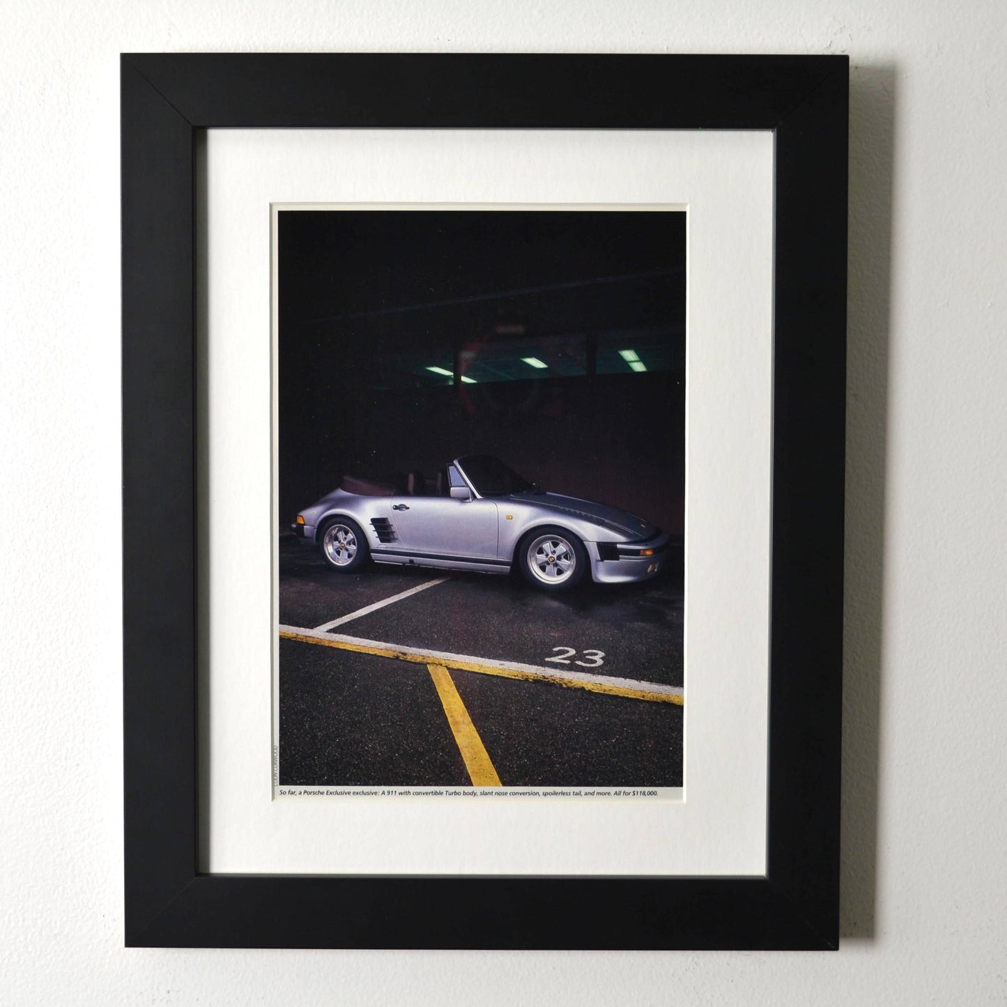 Framed late-1980s Porsche 911 Turbo slantnose cabriolet photo print in silver, parked under garage lights; matte black frame with white mat.