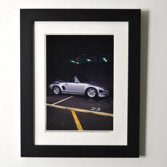 Framed late-1980s Porsche 911 Turbo slantnose cabriolet photo print in silver, parked under garage lights; matte black frame with white mat.