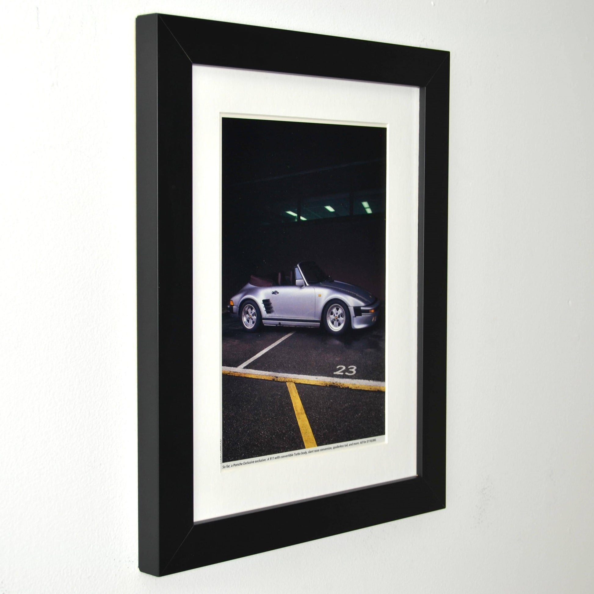 Framed late-1980s Porsche 911 Turbo slantnose cabriolet photo print in silver, parked under garage lights; matte black frame with white mat side view