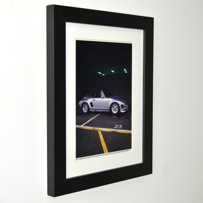 Framed late-1980s Porsche 911 Turbo slantnose cabriolet photo print in silver, parked under garage lights; matte black frame with white mat side view